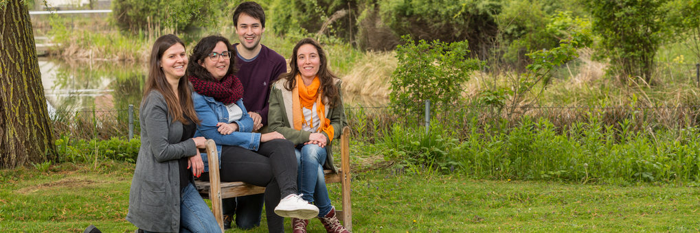 Four PhD students sit outside by the pond and look into the camera.